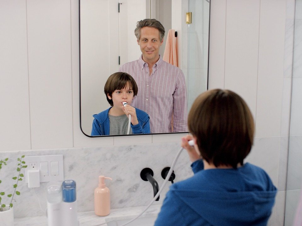 Young boy using a water flosser while his father watches in the mirror during a morning routine